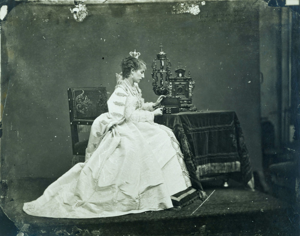 Sarah Bernhardt avec une grande robe blanche assise à une console et avec une boite à bijoux. 
elle joue le rôle de la Reine dans Rue Blas de Victor Hugo.